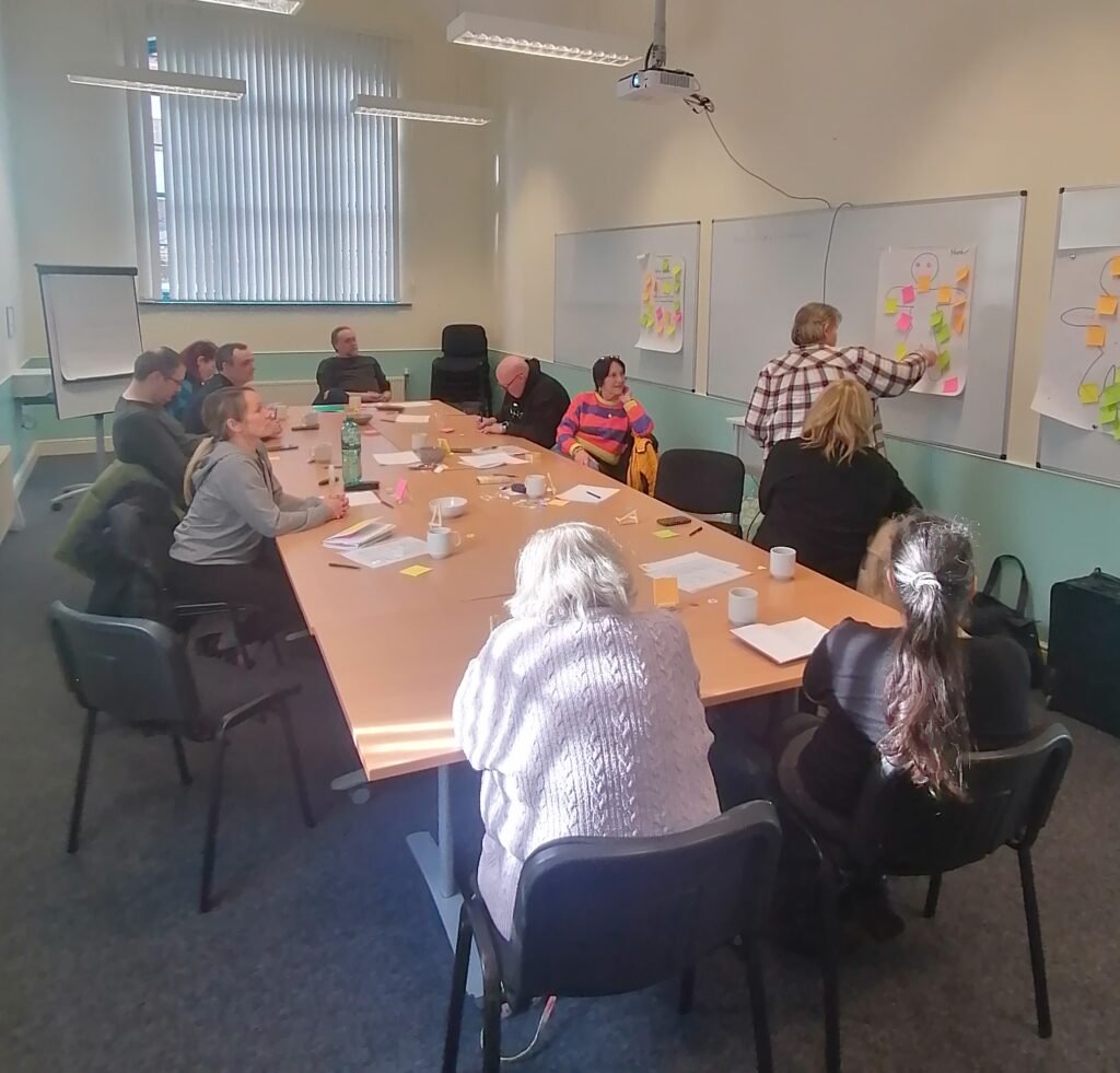 A group of people are seated around a table in a well-lit room, facing a whiteboard. Three large sheets of paper, each featuring the outline of a human figure, are attached to the board. The figures are covered with colourful sticky notes. One person stands at the front with their back to the group, placing sticky notes onto one of the paper figures, suggesting a collaborative workshop session.