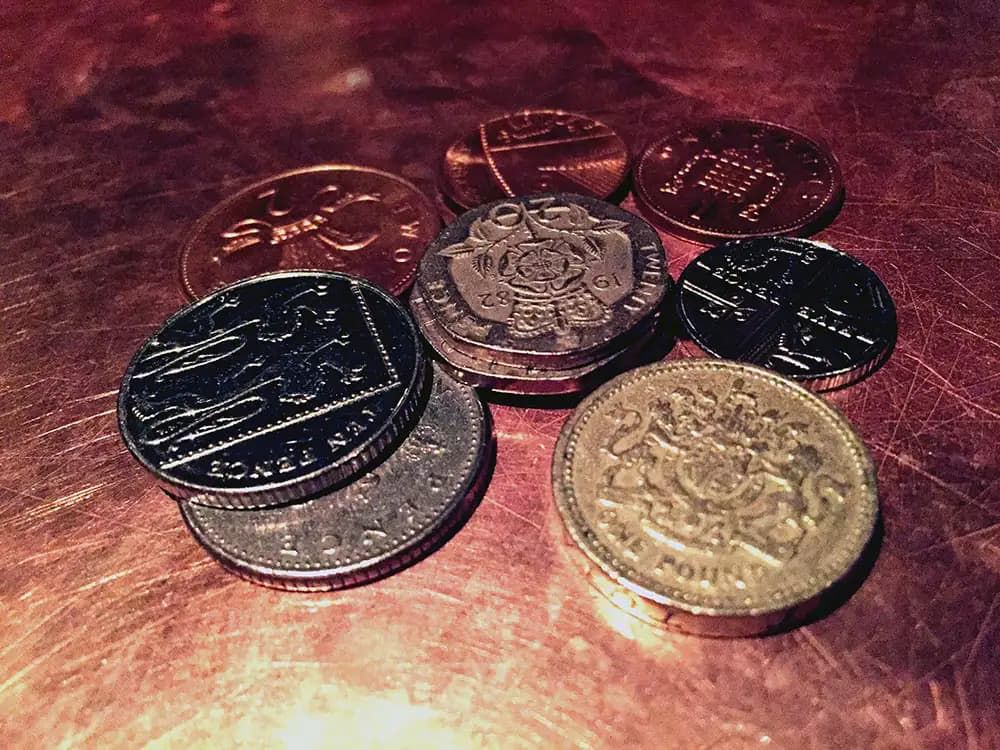 A close up photo of assorted British coins scattered on a table, including a gold-coloured one-pound coin, a silver 20 pence piece, a few 10 pence coins, and several copper coins.