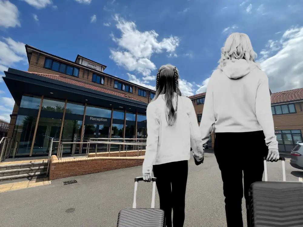 An image of a mother (right) and daughter pulling suitcases along as they walk towards James House. They are wearing light grey tops and black trousers and look 'black and white' against the blue sky.