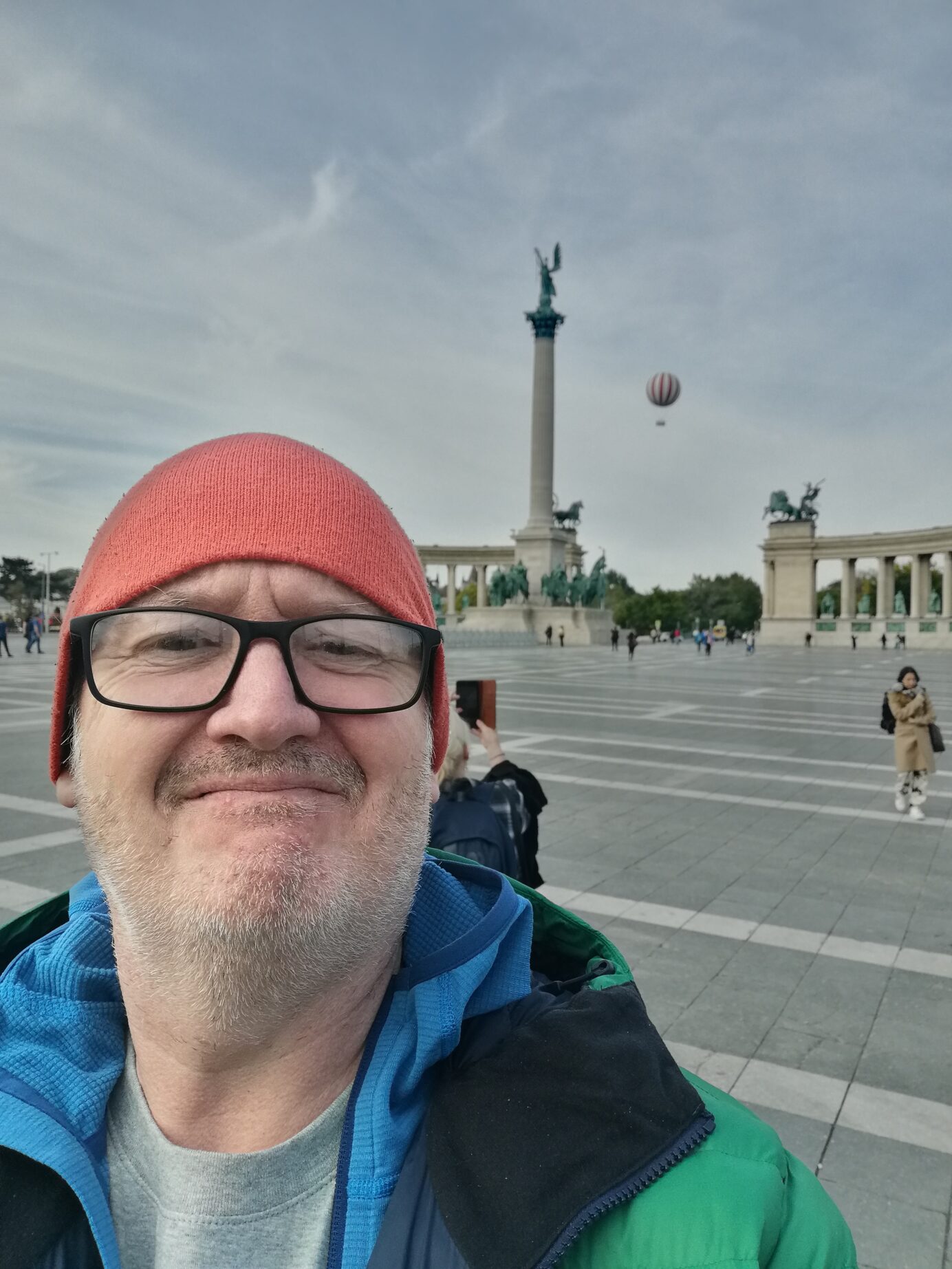 A photo of Miles in Budapest wearing an orange hat and blue coat. There is a hot air balloon in the background and historical monument in the background.