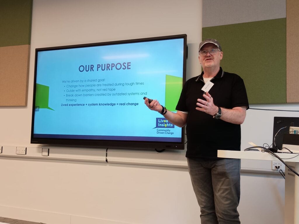 Miles standing on stage presenting at a conference, gesturing toward a PowerPoint slide titled 'Our Purpose' displayed behind him.