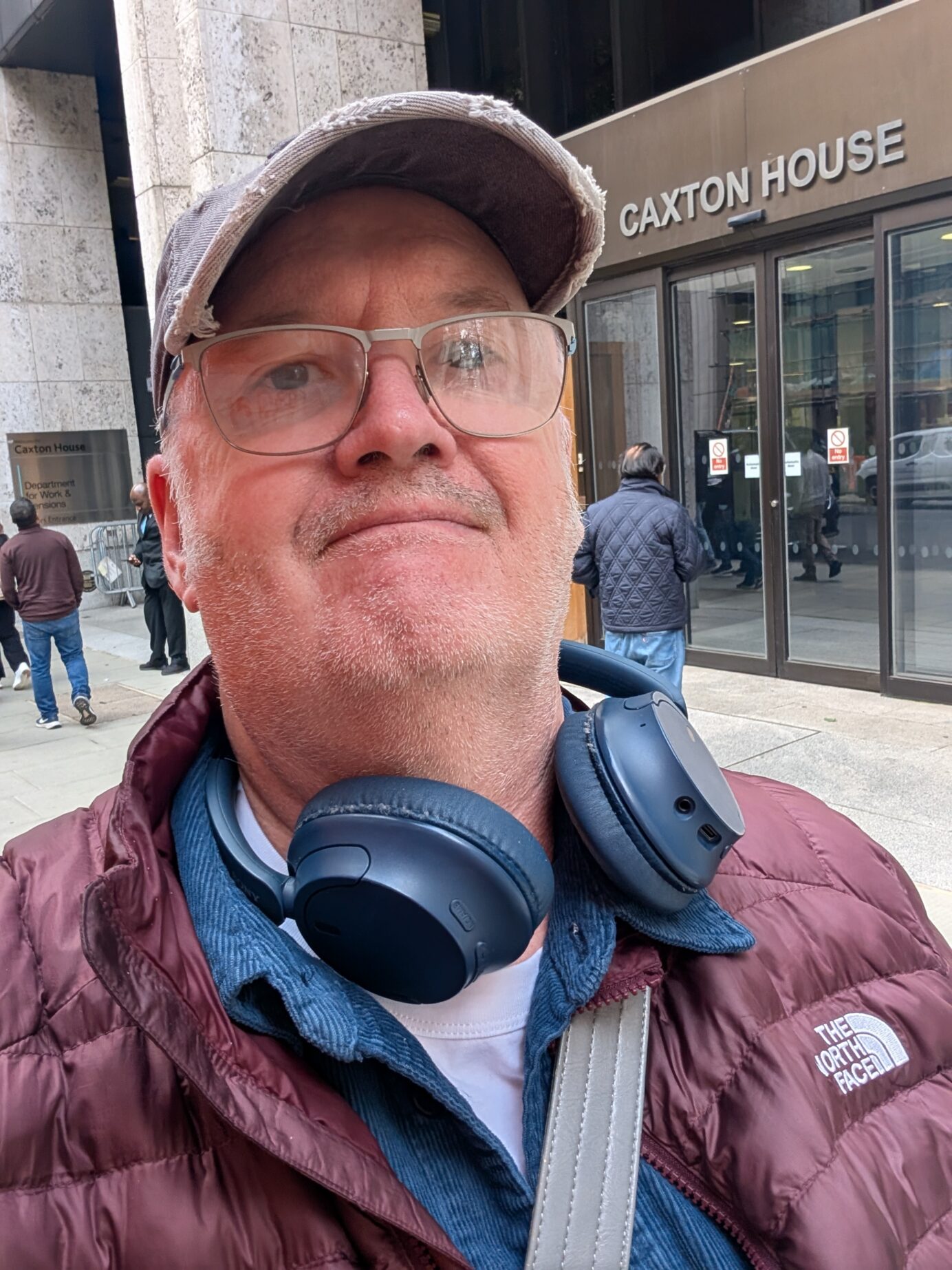 Miles standing outside Caxton House, wearing glasses, blue headphones around his neck, and a purple coat. The Caxton House building is visible in the background.