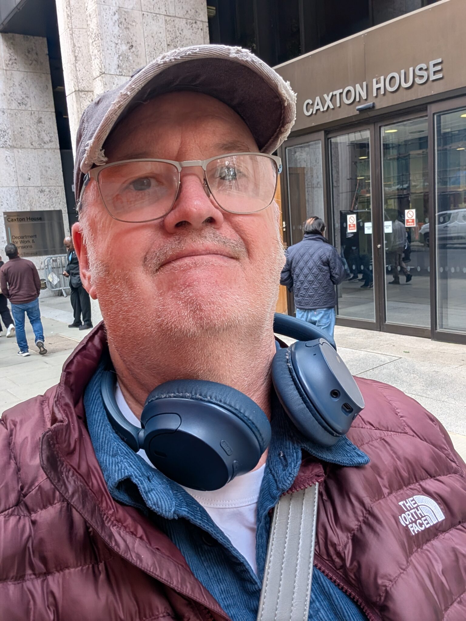 Miles standing outside Caxton House, wearing glasses, blue headphones around his neck, and a purple coat. The Caxton House building is visible in the background.
