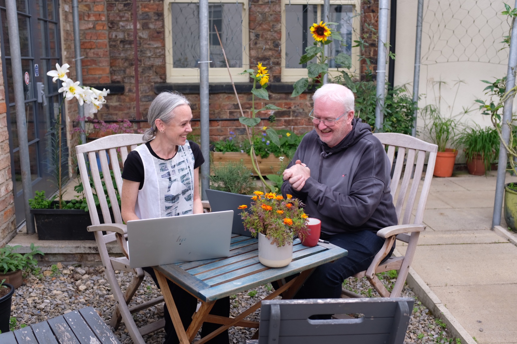 Astrid and Miles sit at a small outdoor table in the garden courtyard at York CVS, looking at a laptop together. Potted flowers and sunflowers surround them.